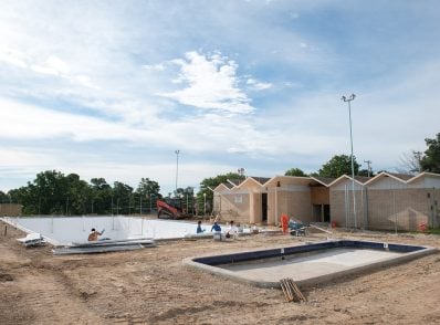 Construction site of a new pool complex under a blue sky. Workers install tiles in empty pools, surrounded by dirt and unfinished buildings.