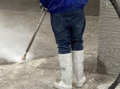 Worker in white boots and a blue jacket power-washes a concrete surface. The scene captures a sense of cleanliness and effort.