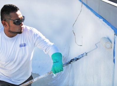 Man painting a pool wall white with a roller, wearing sunglasses, green gloves, and a white shirt. He appears focused on the task.