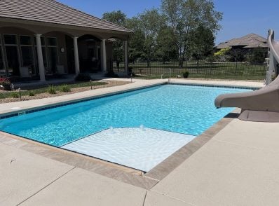 Backyard with a rectangular pool, waterslide, and bubbling fountain. Surrounded by a patio, garden, and trees under a clear blue sky. Tranquil setting.