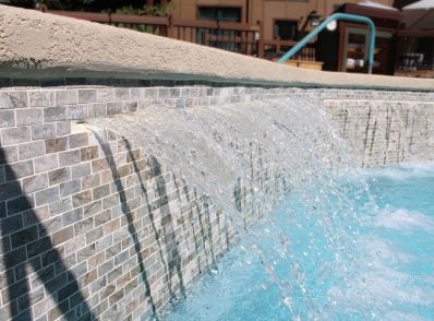 Water cascades over a tiled ledge into a pool, creating a relaxing and refreshing scene. The clear blue water contrasts with the mosaic tiles.