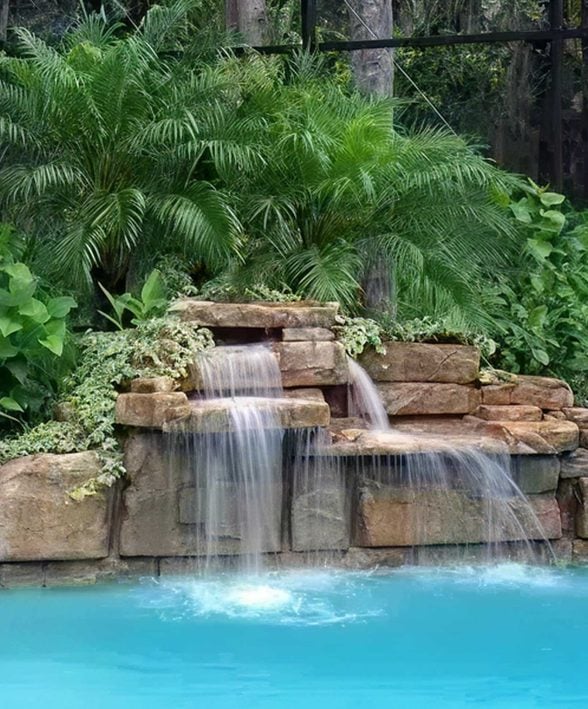 A serene waterfall flows over stacked rocks into a turquoise pool, surrounded by lush green foliage, conveying a tranquil and tropical ambiance.